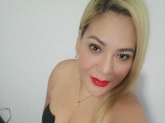 MyaLopez has blonde hair, red lipstick, and a black strapless top, showing her small tits and a gold necklace, with a slight smile. She's looking up at the camera.