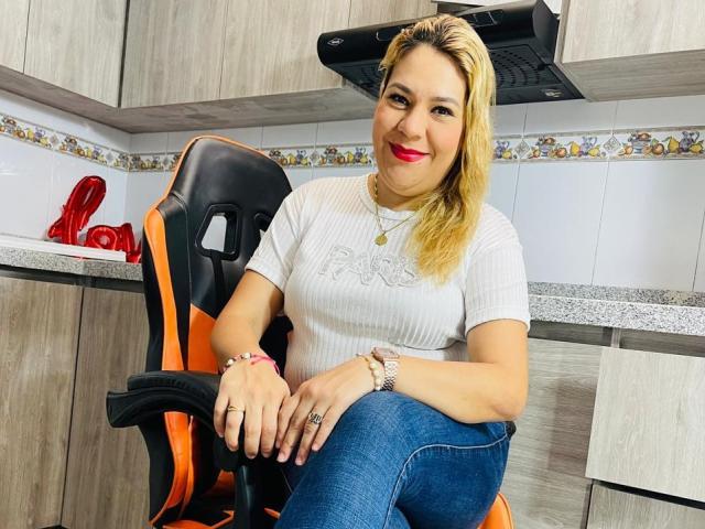 Mya Lopez, met blond haar, rode lippenstift, een wit geribbeld shirt, blauwe jeans, een oranje-zwarte gaming stoel, lachend, in de keuken, met een armband en ring om, zittend, handen op de knieën, borsten zichtbaar..