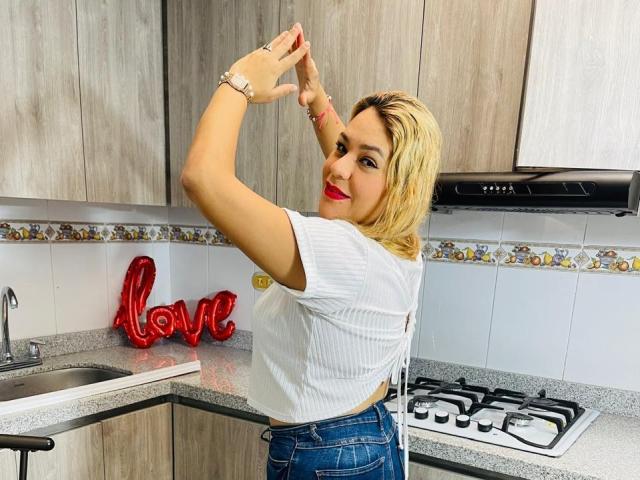 Mya Lopez, con i capelli biondi, una maglietta bianca a coste e jeans blu, sorridente, con le braccia alzate, un insegna al neon rossa con la scritta "amore", una cucina, un lavello in acciaio inossidabile e pensili in legno grigio..