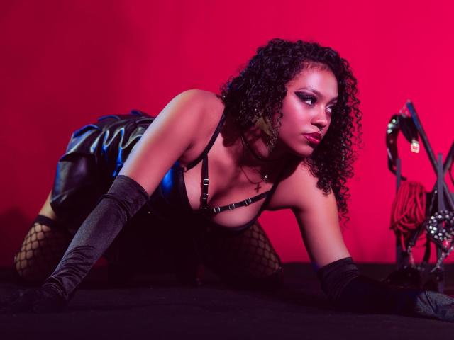 EmilyThonsoon on all fours, wearing black leather dress, fishnet stockings, and gray elbow gloves, with curly hair, black harness, and pink eye makeup, looking to the side.