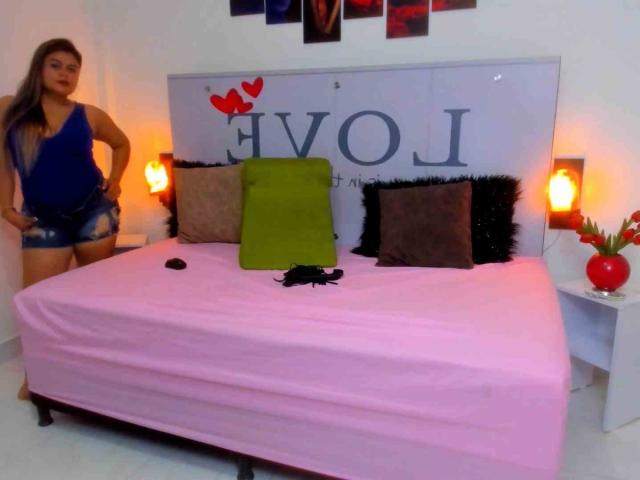 CaandyFoox stands by a pink-covered bed with green and brown pillows, black feathers, red heart decor, and orange lamps. She wears a blue top and denim shorts.