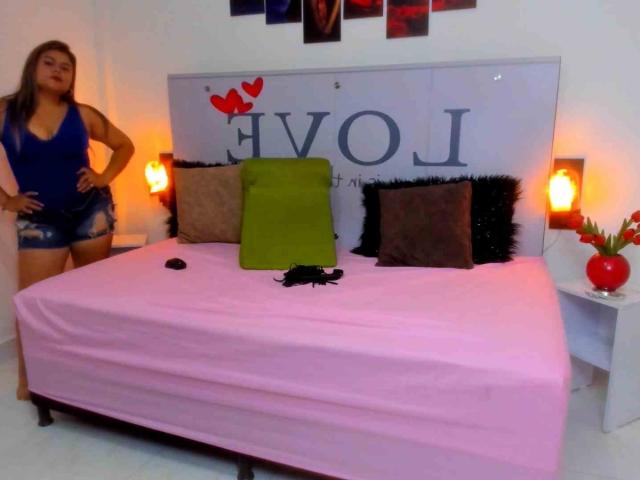 CaandyFoox stands with hands on hips, wearing a blue top and denim shorts, next to a pink-covered bed with green and brown pillows, and two lit lamps.