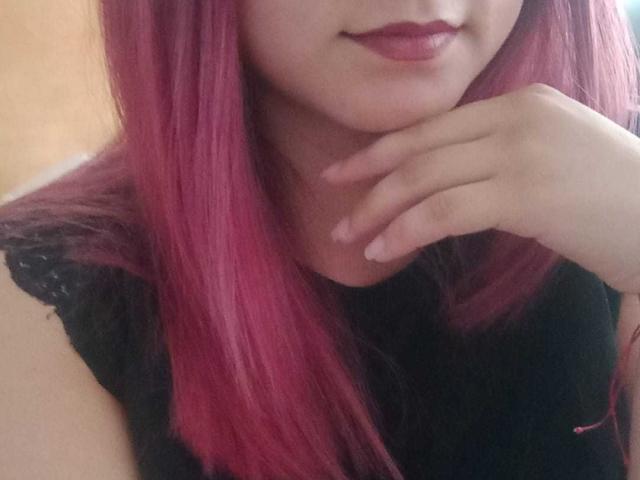 LiaJonnesX with pink hair, black lace top, and red lipstick, resting her chin on her hand, showing off her cute, small tits. Her fingers are gently touching her neck.
