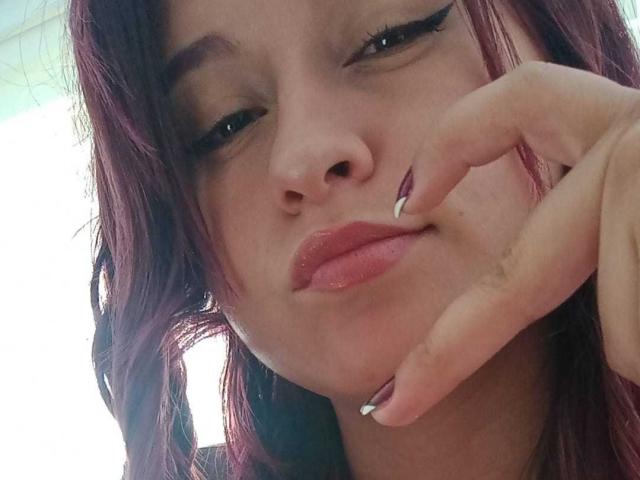 LiaJonnesX has purple-dyed hair, glossy pink lips, and is touching her chin with her right hand, showing off her manicured nails. Her eyes are half-closed in a slight smile.