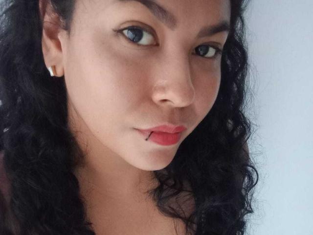 DanielaBebe hat ein Gesicht mit lockigem, schwarzem Haar, hellbrauner Haut, blauen Augen und einem kleinen schwarzen Piercing an ihrer Unterlippe. Sie trägt einen goldenen Ohrring..