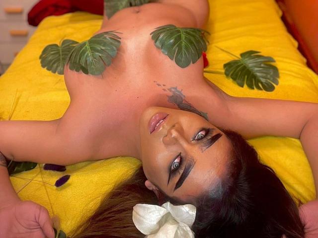 EmpressiveValerie is lying topless on a yellow bed, with large green leaves covering her tits, arms above her head, and a white flower in her brown hair.