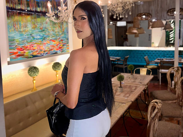FallonRogers with long black hair, wearing a black strapless top and white pants, holding a black bag, standing in a restaurant with a colorful painting and chandeliers.