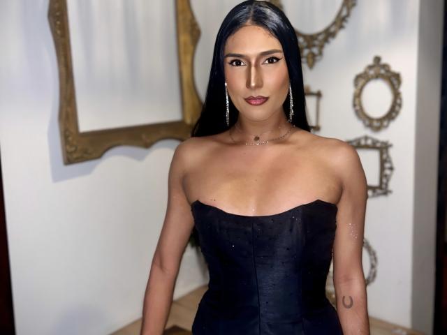 FallonRogers with long black hair, wearing a strapless black corset, showing small tits, dark lipstick, and earrings, standing in a room with gold-framed mirrors.