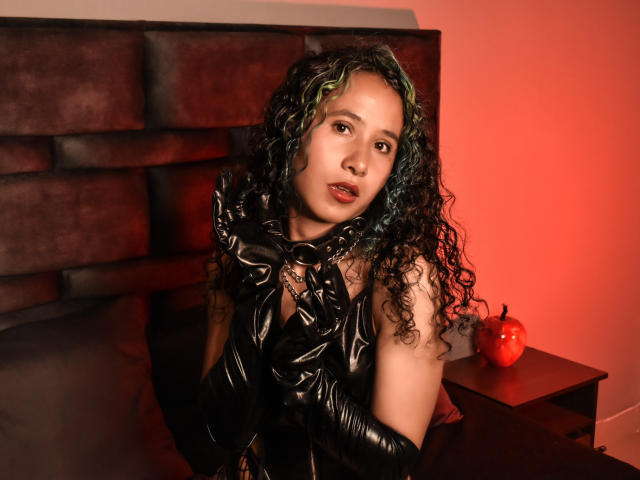 JessyConnors with curly green and black hair, wearing black latex gloves and dress, sitting on bed, red light, apple on nightstand, looking at camera with slightly open mouth.
