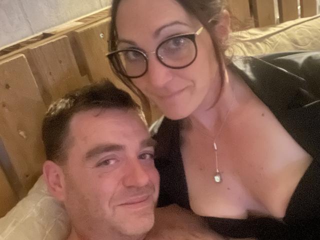 TifolyLaurent, avec des lunettes, des cheveux foncés et un collier, et un homme torse nu avec des cheveux bruns courts, souriant à la caméra, dans une chambre en bois chaleureuse. Seins visibles..