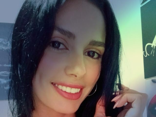AnaisHilton has dark black hair, brown eyes, and a cute smile with red lipstick. She's touching her hair with a ring on her finger, looking sexy.