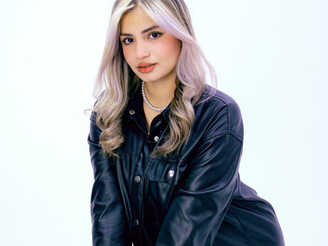AlexaThoms has blonde, wavy hair, wearing a black leather jacket, pearl necklace, and subtle makeup with pink lips, leaning slightly forward with a cute, confident expression.