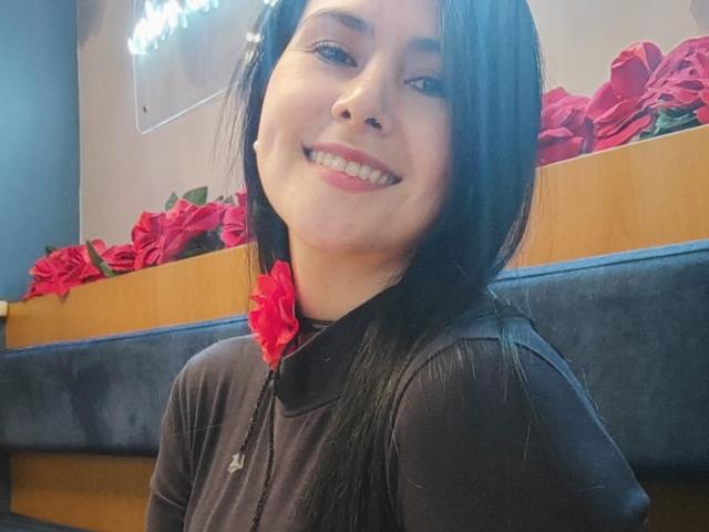 AlmaSil with long black hair, smiling, wearing a black zip-up shirt with a red rose on the chest, showing a nose ring and white teeth. Red roses in background.