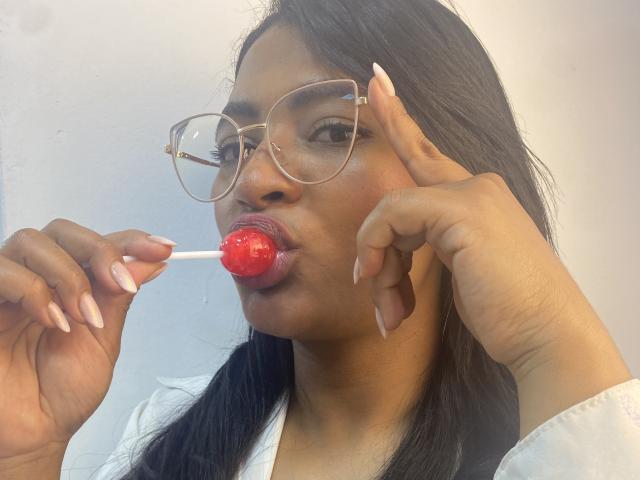 CameronJones with long black hair, wearing glasses, sucking a red lollipop, holding it with her right hand, showing long pink nails, wearing a white shirt.
