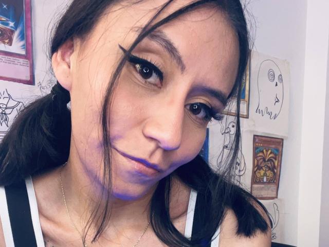 EviieGothel has dark, wavy hair, thick black eyeliner, and a slight purple bruise under her right eye. She's wearing a white and black top, with a subtle smile.