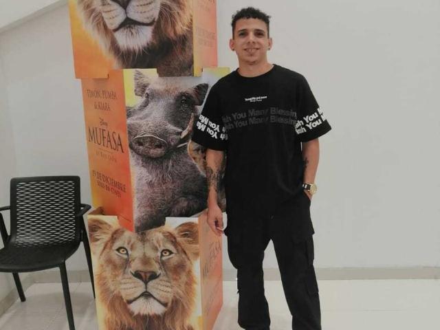 HottLoverGuy69 stands in black clothes, hands in pockets, tattoos visible, in front of animal posters with lion and warthog faces. Black chair on left. Short curly hair. Smiling.