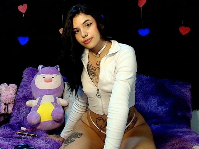 GuadalupeClak mit dunklem Haar, einem weißen Reißverschluss-Oberteil und einem pinken String, sitzt auf einem violetten Fell, zeigt Tattoos und hält ein violettes Plüschtier, im Hintergrund sind pinke und blaue Herzen zu sehen..