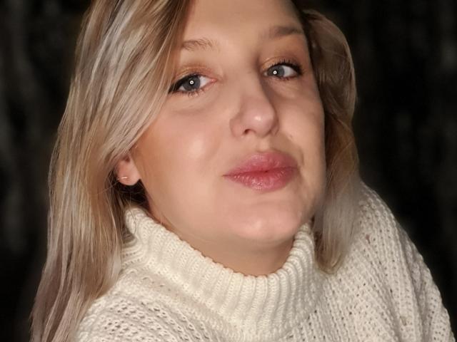 MayaTrainor with blonde hair, wearing a white turtleneck, has pink lips and a slight smile, her face is centered, showing her nose and eyes, with a soft, warm light.
