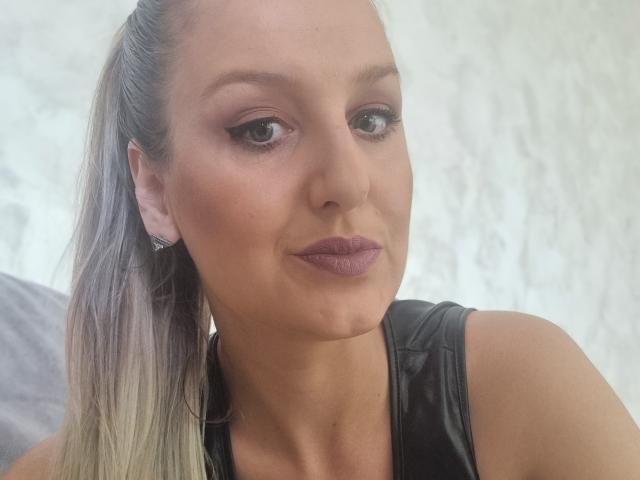 MayaTrainor has blonde ponytail, brown eyes, subtle makeup, mauve lipstick, black leather top, and a slight smirk. Her tits are small, and she's wearing diamond earrings.