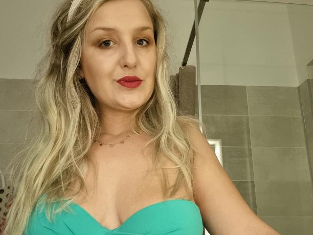 MayaTrainor with blonde hair, red lipstick, green strapless top, showing her tits, standing in a bathroom, looking at the camera, wearing a delicate necklace.