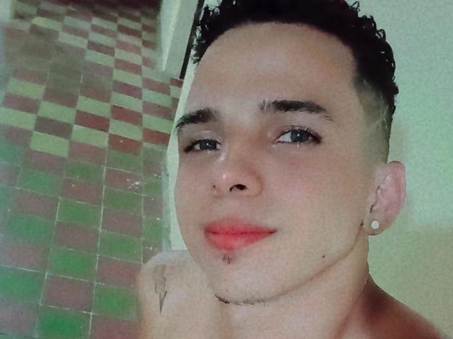 HottLoverGuy69 with short curly black hair, light skin, small nose, red lipstick, ear piercings, subtle tattoo, and a slight mustache, looking at the camera with a slight smile.
