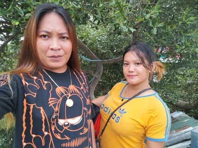 EXOTICPINAY with long brown hair in a black cat-print shirt and EXOTICPINAY with tied-up hair in a yellow Adidas shirt standing outdoors, both looking at the camera.