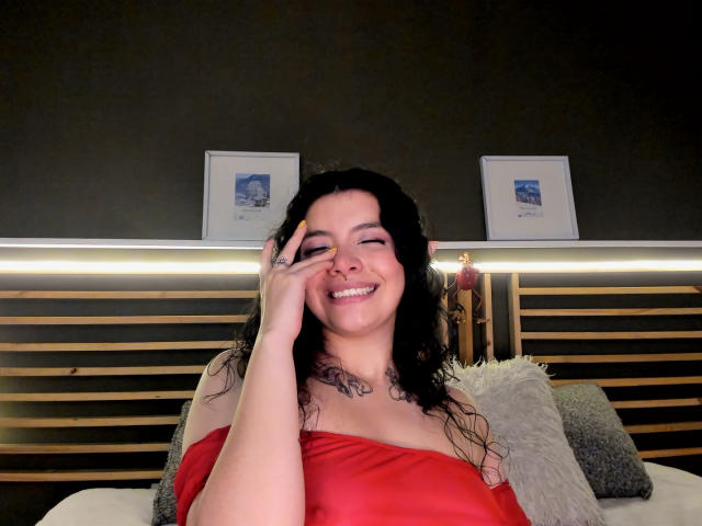 LilibethHot69, con cabello negro rizado, sonriendo, vestida con una blusa roja con un hombro descubierto, tocándose la mejilla, con un tatuaje visible en el cuello, sentada en una cama con listones de madera..