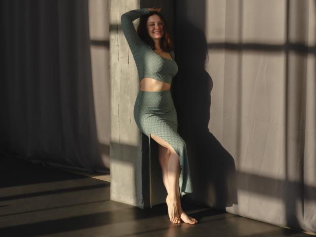 Barbara Stily, pieds nus, portant un crop top vert matelassé et une jupe, bras levé, souriante, debout contre un pilier en béton, la lumière du soleil projetant des ombres, montrant ses seins et ses fesses sur la photo..