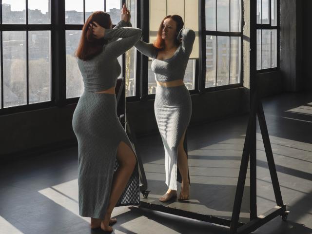 BarbaraStily with red hair, gray knit crop top and skirt, showing ass and tits, standing in front of mirror, raising arms, sunlight, reflection, wooden floor, black frame.