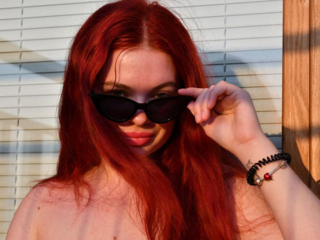 CarolinaDiaa with red hair, black sunglasses, and a black bead bracelet, touching her sunglasses, showing her pink lipstick and a slight smile, with her tits partially visible.