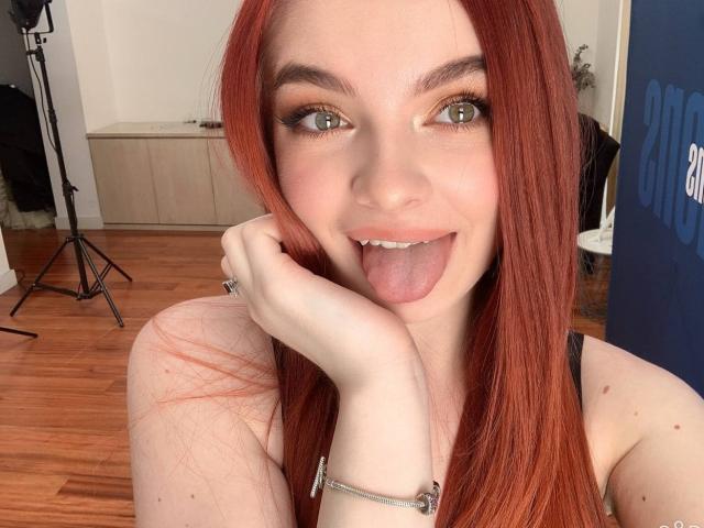 CarolinaDiaa with red hair, green eyes, and fair skin, sticking out her tongue, hand on cheek, wearing a black top, wooden floor, and a silver bracelet.