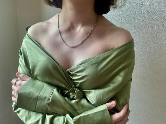 Steisii's green shirt is off-shoulder, showing her tits and a silver chain necklace. She's crossing her arms, her hands touching her elbows, with curly brown hair.