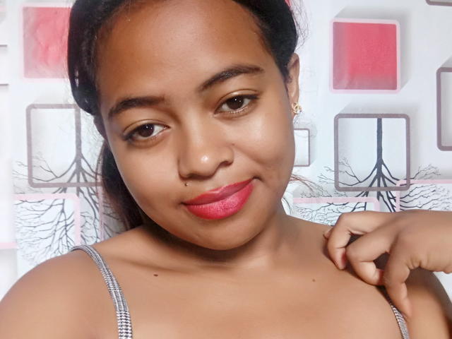 CiciahBella with dark brown skin, red lipstick, small nose piercing, black hair in a ponytail, wearing a gray spaghetti strap top, touching her shoulder, smiling subtly.