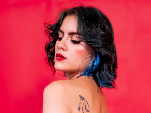 SamHazell with shoulder tattoo, black wavy hair with blue tips, red lipstick, nose ring, and large hoop earrings, against a bright red background.
