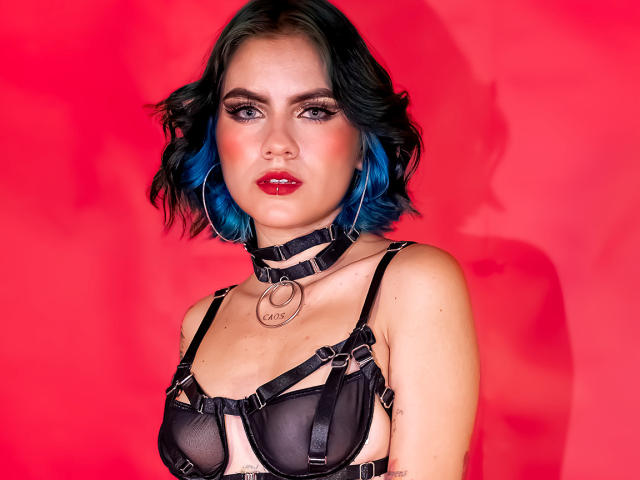 SamHazell with blue-tipped black hair, wearing a black harness, choker with ring, and red lipstick, showing tattoos, has a serious expression against a bright red background.