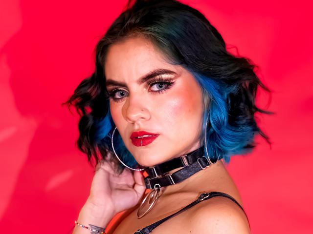 SamHazell with blue and black hair, red lipstick, hoop earrings, black collar with ring, looking at camera, hand near face, red background, showing tits slightly in black top.