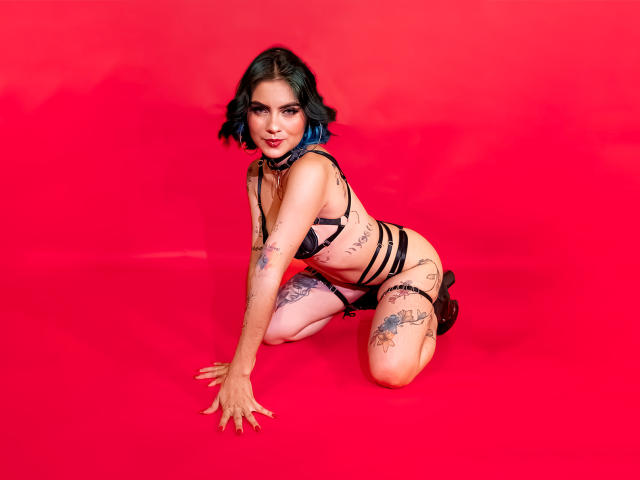 SamHazell is on her knees, wearing black strappy lingerie, with blue-dyed hair, tattoos, and black boots, against a bright red background. Her tits are small, and she's sexy.