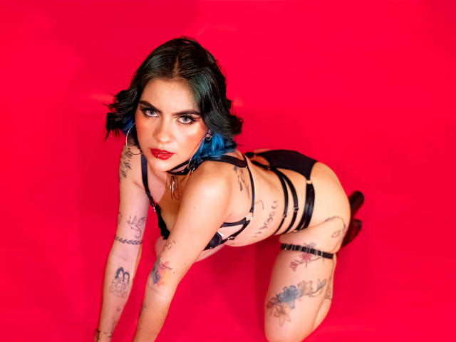 SamHazell with blue hair, red lipstick, black strappy lingerie, tattoos, and a seductive gaze on all fours against a bright red background. She's got a sexy, curvy ass.