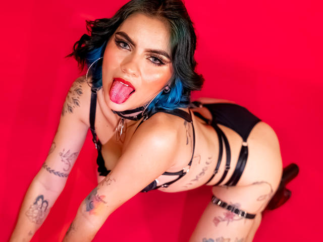 SamHazell with blue hair, black strappy lingerie, tattoos, and tongue out, on red background, showing her ass and looking up. She's wearing a black choker and garter.