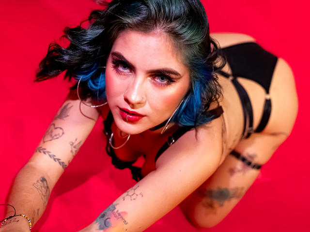 SamHazell with blue-dyed hair, black strappy lingerie, tattoos on arms and thighs, kneeling on red, looking up with red lipstick and nose ring, showing her ass.