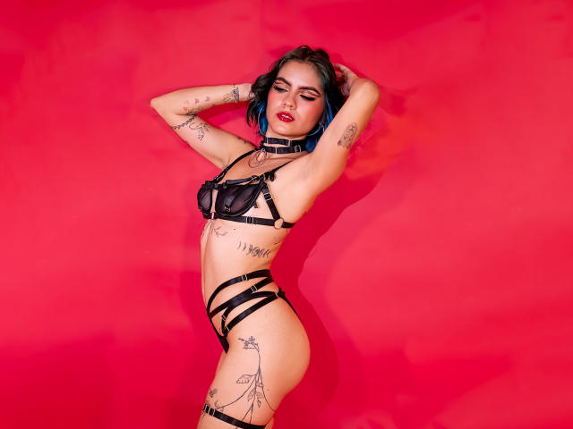 SamHazell with blue-dyed hair, wearing black strappy lingerie, tattoos on arms and thighs, red background, arms raised, eyes closed, red lipstick, looking sexy, standing, confident.