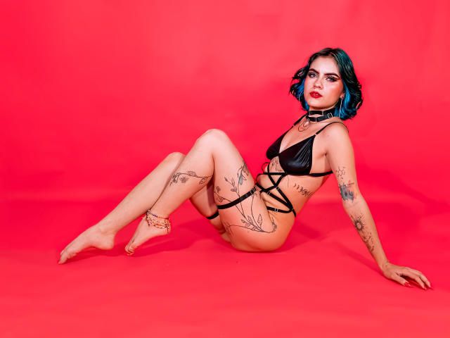 SamHazell with blue hair, black strappy bikini, tattoos, red background, sitting with legs bent, arms behind, looking at camera, red lipstick, bare feet, black choker.