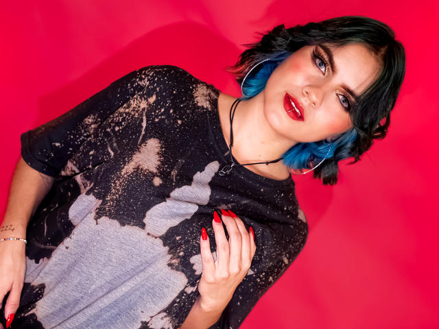 SamHazell with blue hair, red lipstick, and red nails, wearing a black, splattered t-shirt, touching chest, against a bright red background. She has a tattoo on her right hand.