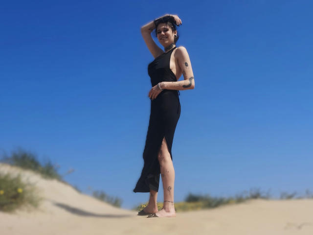 SpiceDevil-hot in a black, sleeveless, high-slit dress, standing barefoot on sand, arm raised, tattoos visible, clear blue sky, short dark hair, smiling, summer vibe.