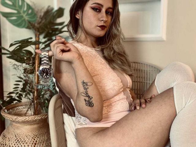 LiaJonnesX with blonde hair, wearing lace pink bodysuit, white thigh-highs, holding wristwatch, showing cursive tattoo on left arm, sitting with legs spread, showing her pussy.