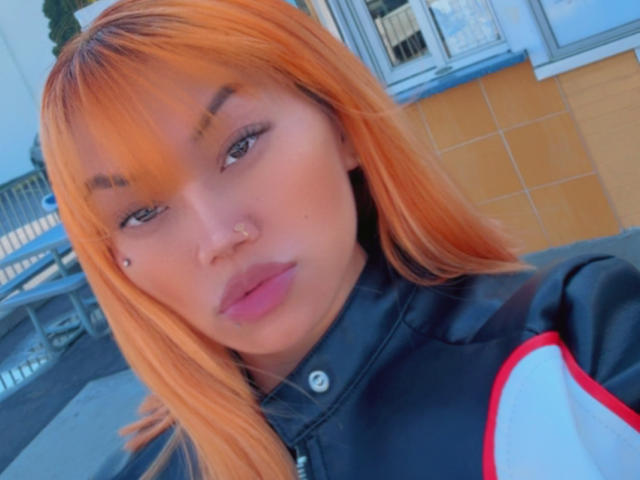 ForeignMaia with orange hair, black jacket, pink lipstick, nose ring, and a small beauty mark on her cheek, looking at the camera, showing her tits slightly in the frame.