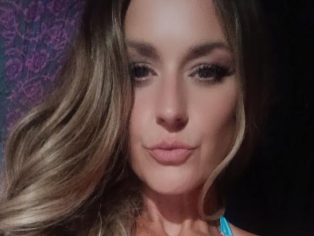 SugxrLips has blonde, wavy hair, full eyebrows, and pink lipstick. She's looking down with a slight smile, showing off her pouty ass-kissing lips. Her eyes are half-open.