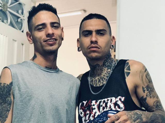 Two BryannThdenverHot guys with tattoos, short dark hair, and ear gauges, wearing sleeveless shirts, standing close, one in gray, one in black "Stars & Stripes" shirt.