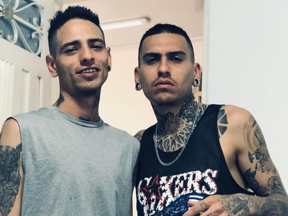 Two BryannThdenverHot guys with tattoos, short dark hair, and ear gauges, wearing sleeveless shirts, standing close, one in gray, one in black "Stars & Stripes" shirt.
