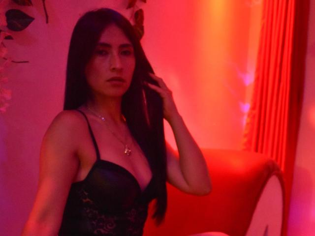 JainaRogers with long black hair, wearing a black lace bra, red lighting, holding her hair, small cross necklace, looking serious, red curtains in the background.